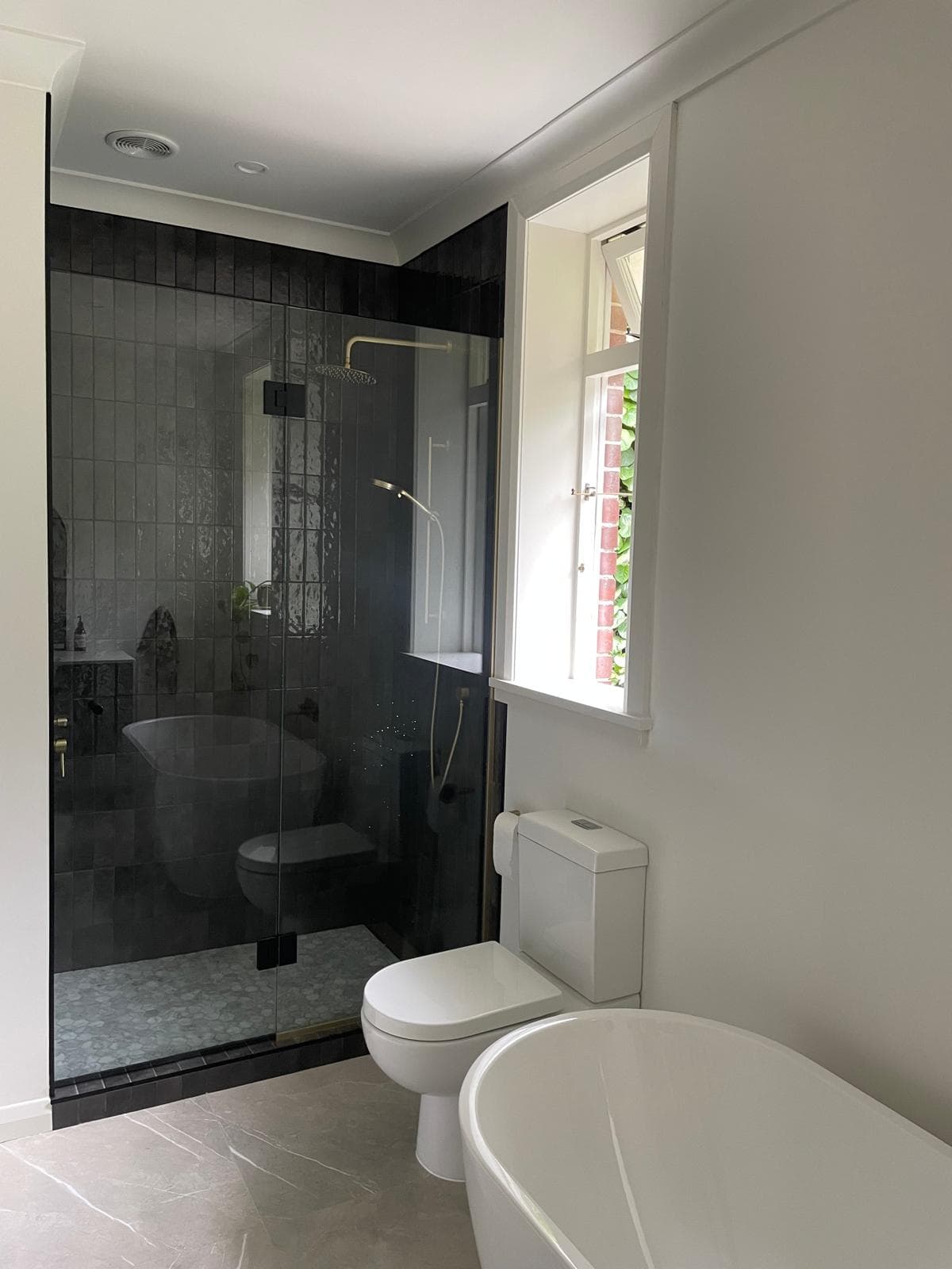 Renovated bathroom with a black-tiled walk-in shower, brass rain head, freestanding white bath and marble-look tiled floor