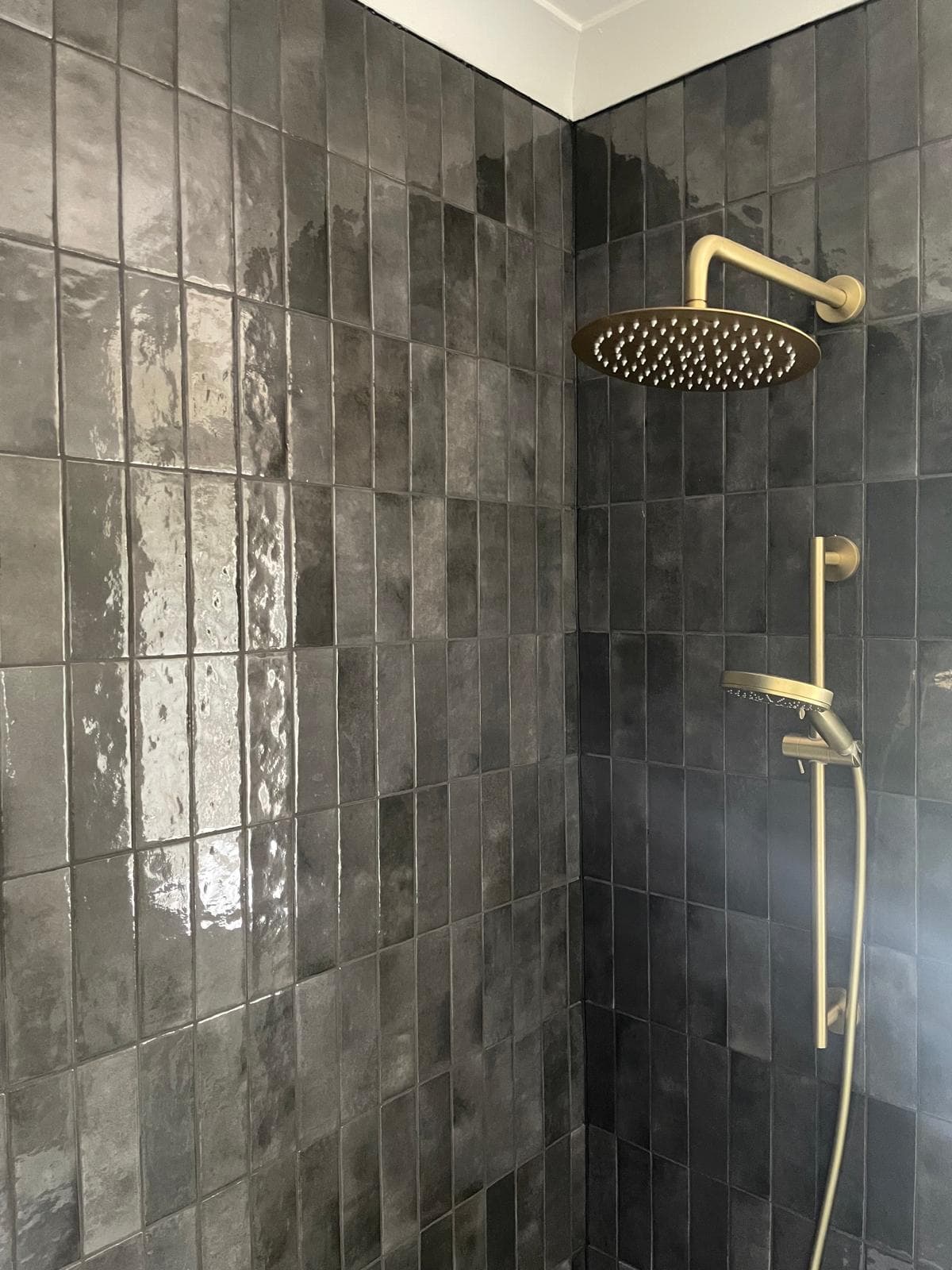 Close-up of a brass rain shower head and slide-rail handset against glossy charcoal wall tiles