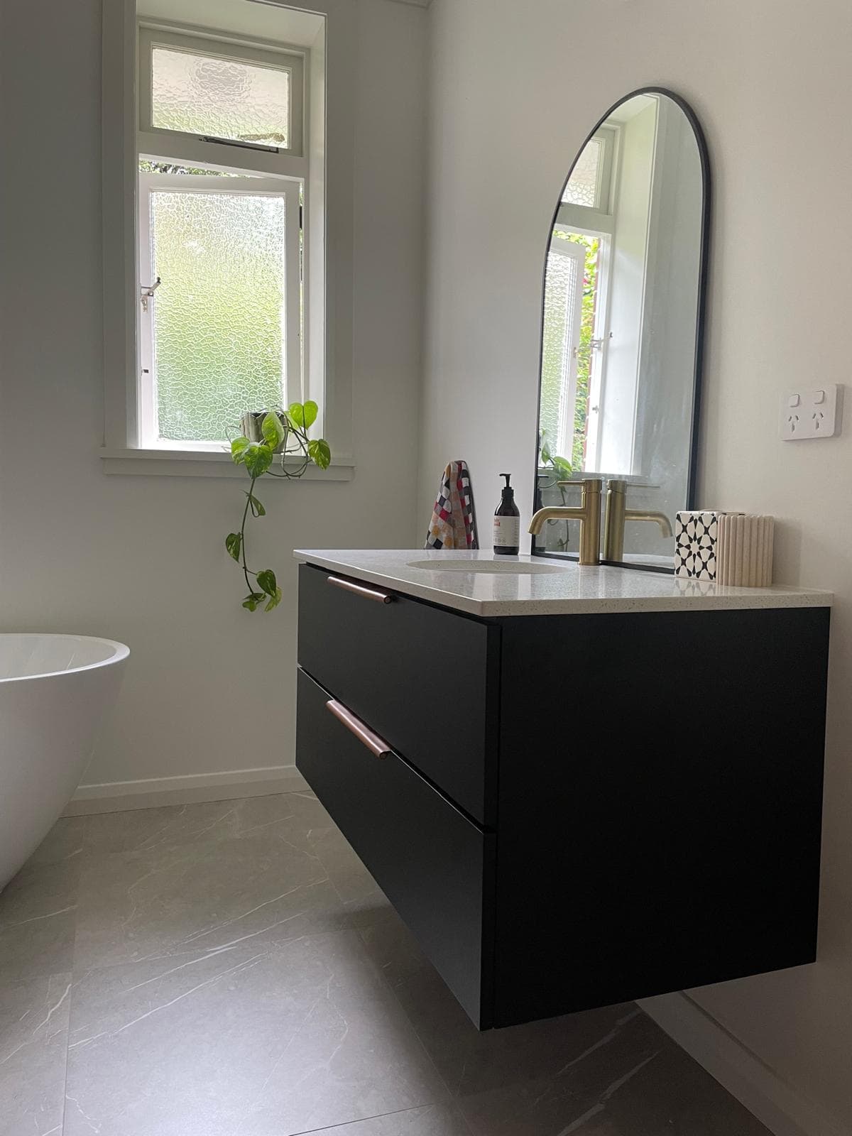 Black floating vanity with a speckled stone top, brass tapware and arched mirror beside a freestanding bath