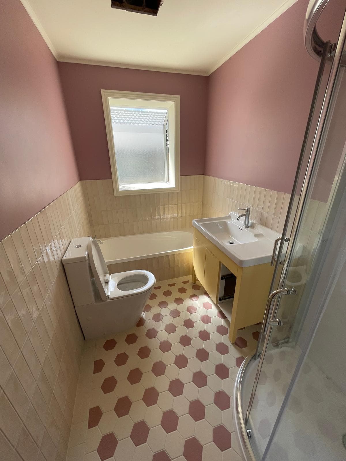 Renovated bathroom with pink walls, cream tiled wainscot, bath, yellow timber vanity and corner shower over a hexagon tiled floor