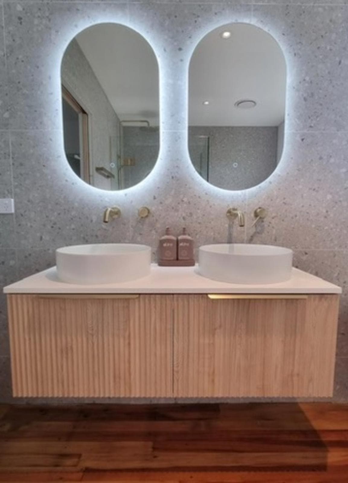 Double timber-fluted floating vanity with twin round basins, brass wall-mounted taps and backlit oval mirrors on grey terrazzo tile