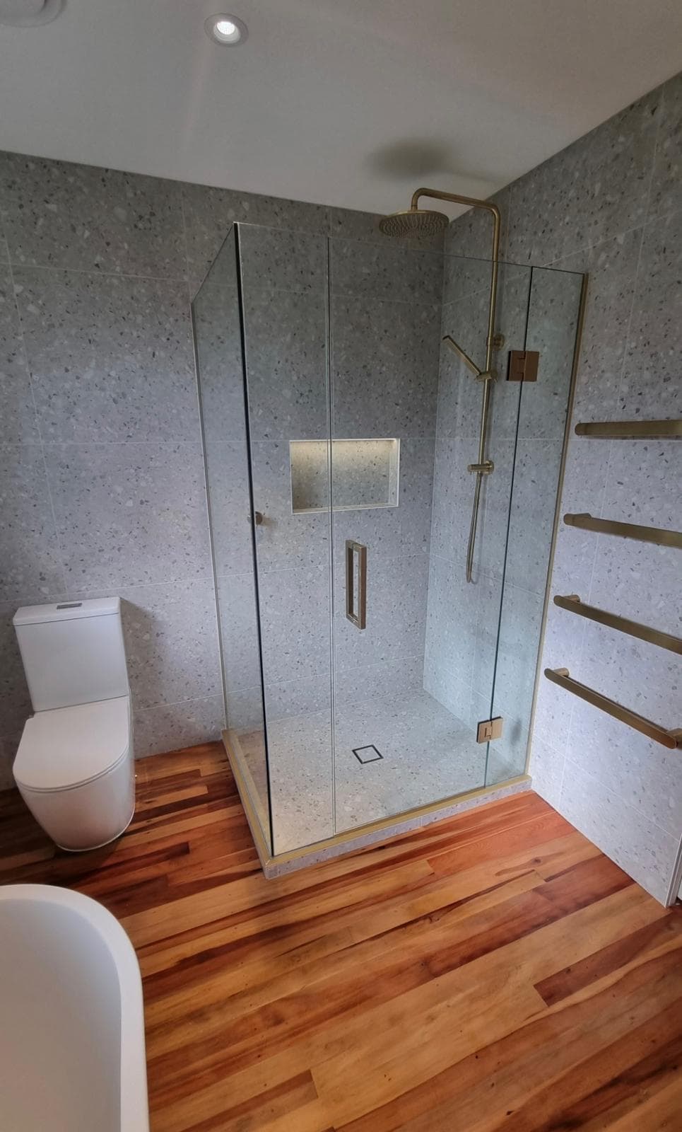Frameless glass corner shower with brass rain head and heated towel rails on grey terrazzo walls above a native timber floor