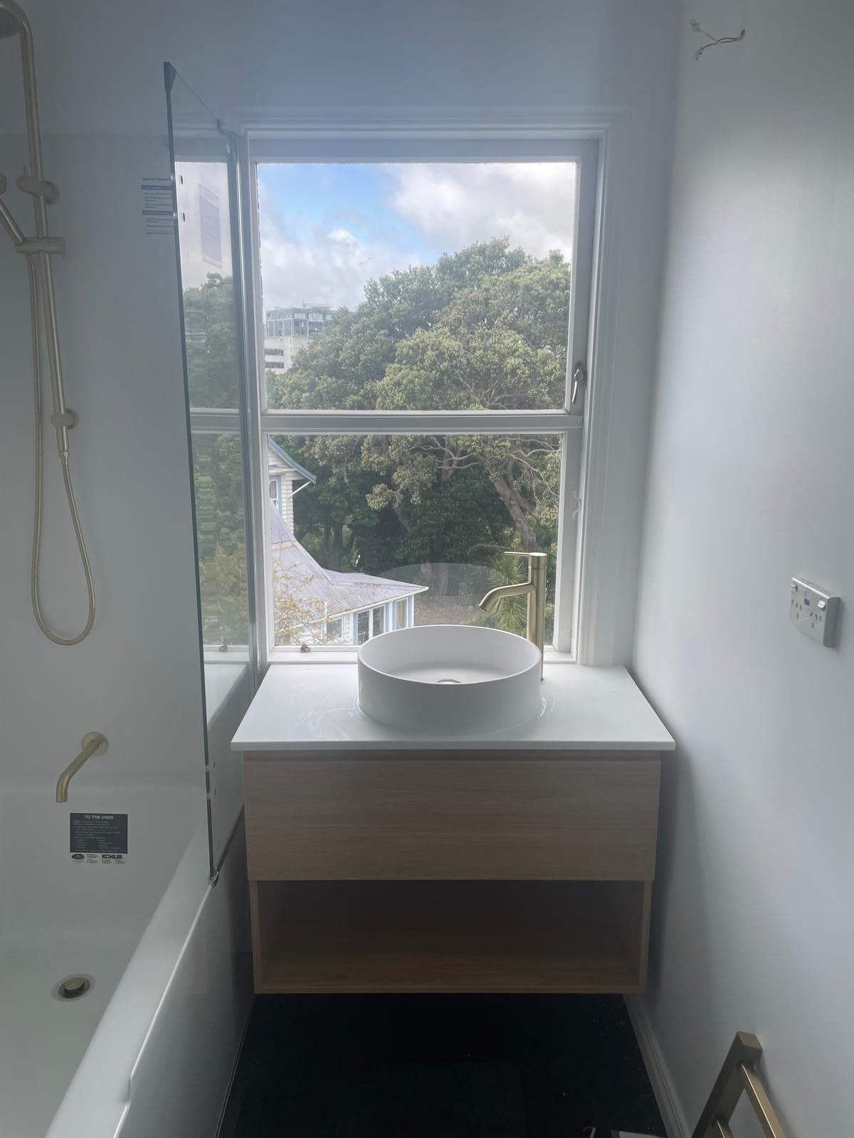 Timber vanity with round countertop basin and brass tall mixer in front of a sash window, next to a bath with brass shower over