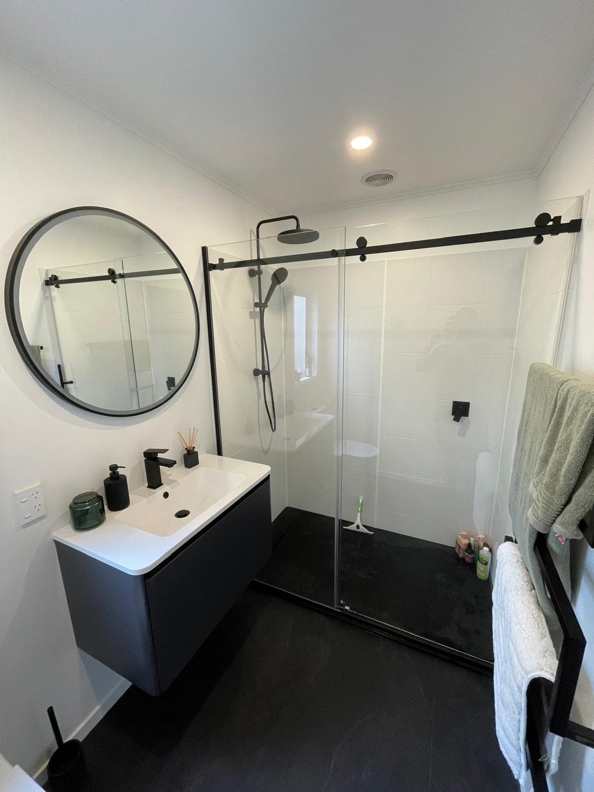 Black and white ensuite with matte black tapware, charcoal floating vanity, round black mirror and sliding-door shower on a black tiled floor