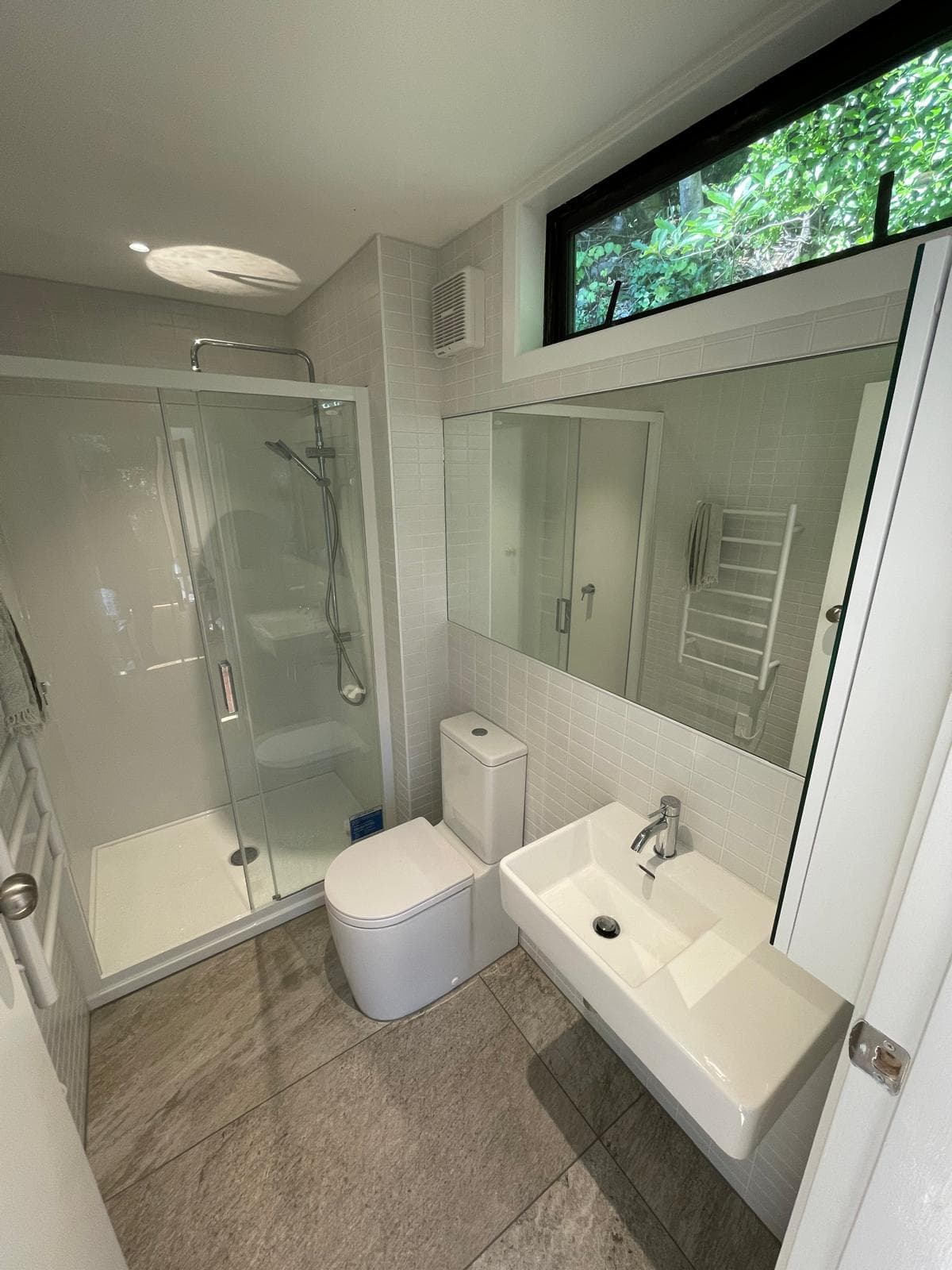 Compact white ensuite with sliding-door shower, wall-hung basin and toilet under a bush-outlook window