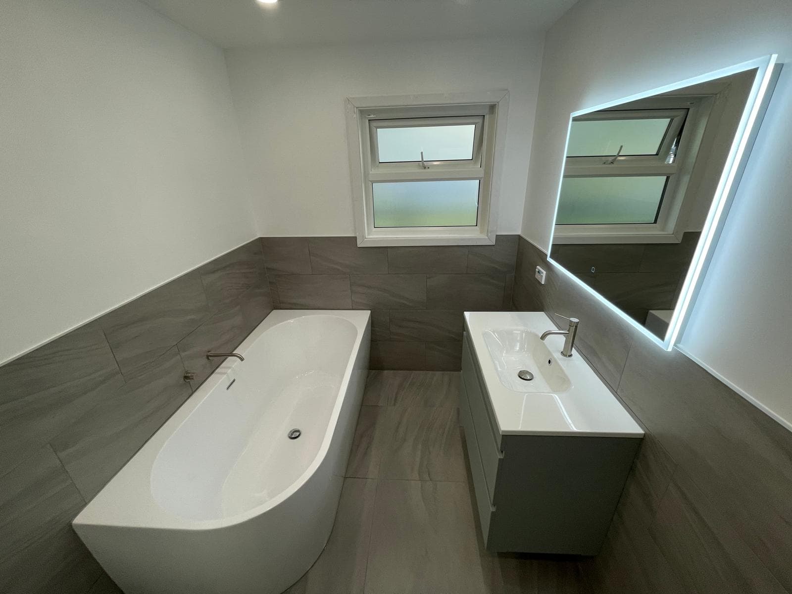 Back-to-wall freestanding bath and grey vanity with LED mirror in a bathroom tiled in grey stone-look tiles