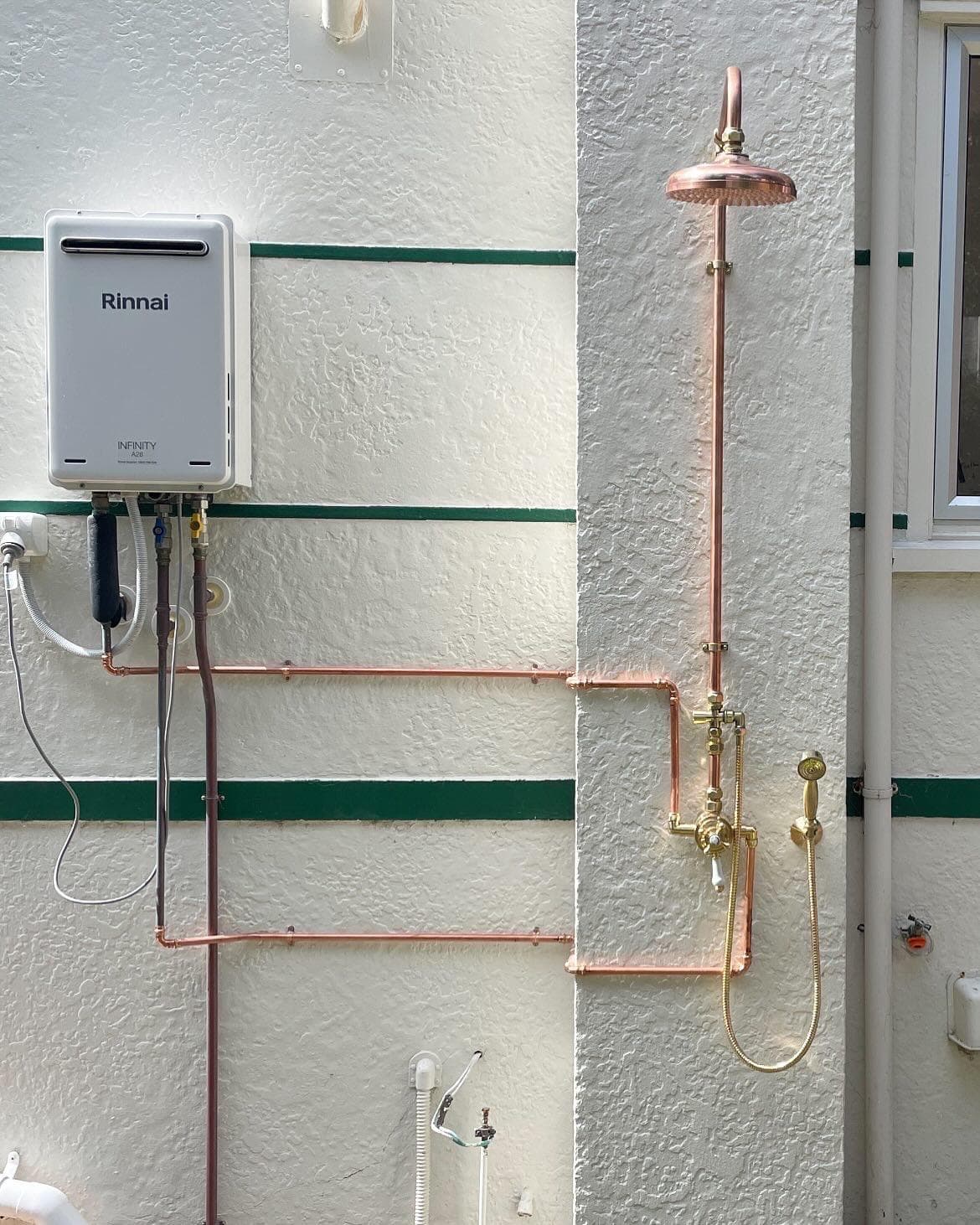 Outdoor copper shower with brass mixer and handset beside a Rinnai Infinity gas water heater on a stucco wall