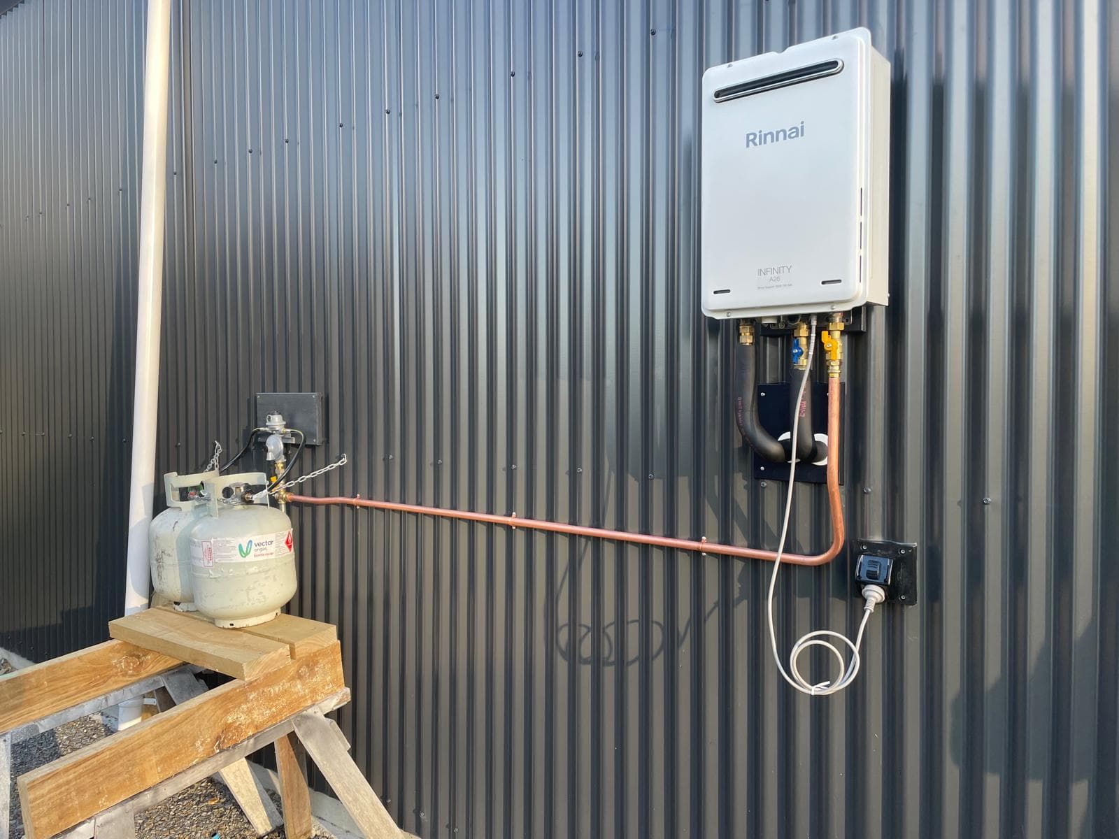 Rinnai Infinity continuous-flow gas water heater installed on a corrugated wall with copper gas line to twin LPG bottles