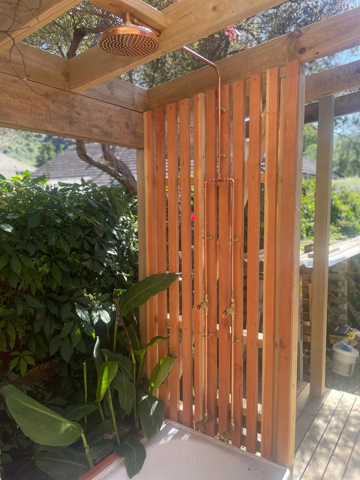 Outdoor shower with copper rain head and exposed copper pipework with brass taps on a timber slat screen, above an outdoor bath